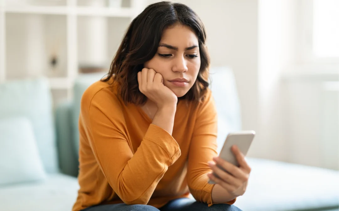 A young woman sitting on a couch looking down at her phone with a worried expression