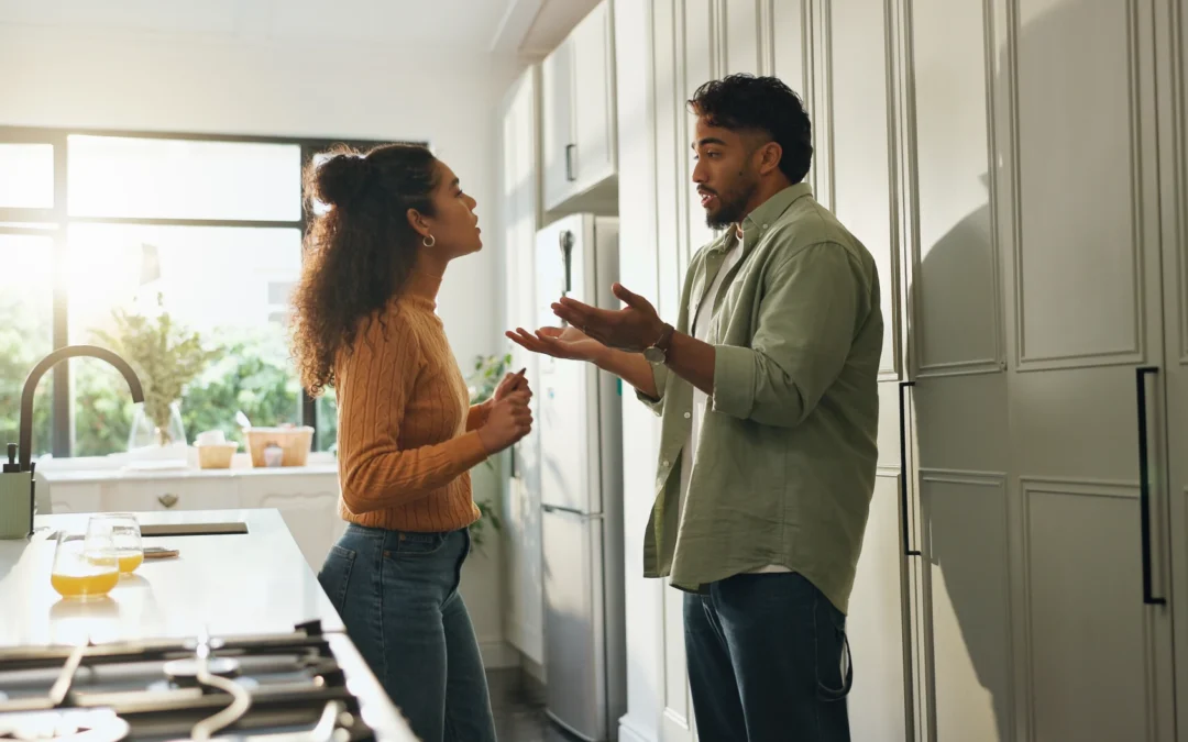 A man and a woman standing in a kitchen having a tense conversation, with the woman’s arms crossed