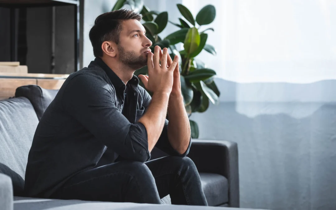 A tired-looking man sitting on a couch at home, resting his head against his hand