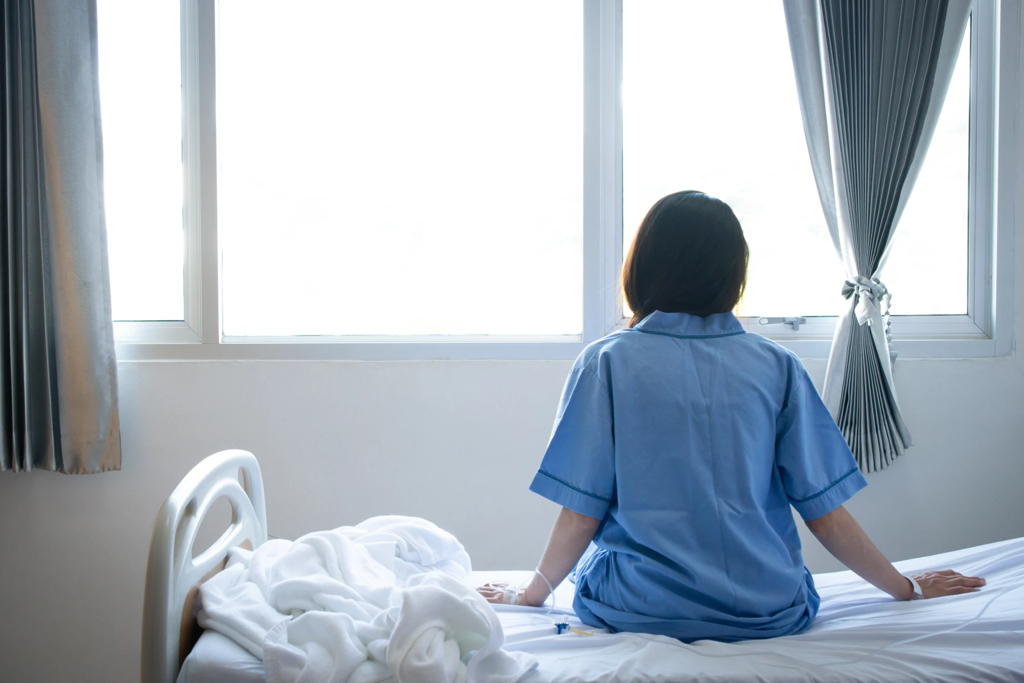 A person resting in a hospital bed near a bright window, with a blanket over their legs