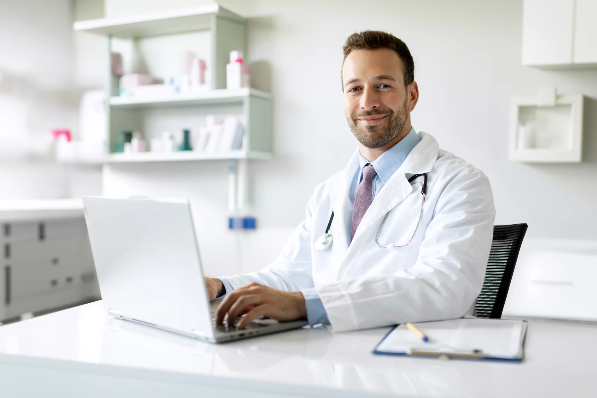 A doctor in a white coat sitting at a desk in a bright office, looking at a laptop with a calm expression