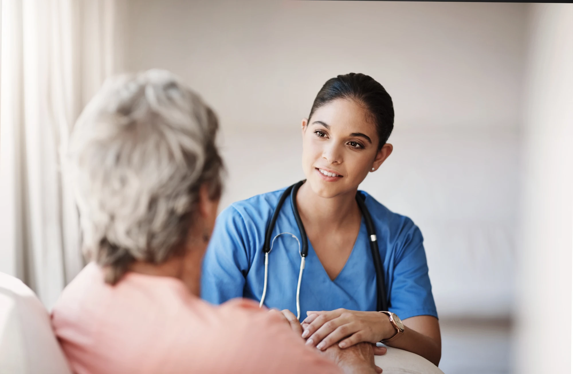 A nurse in scrubs sitting beside a woman on a couch, talking with her in a calm, bright room