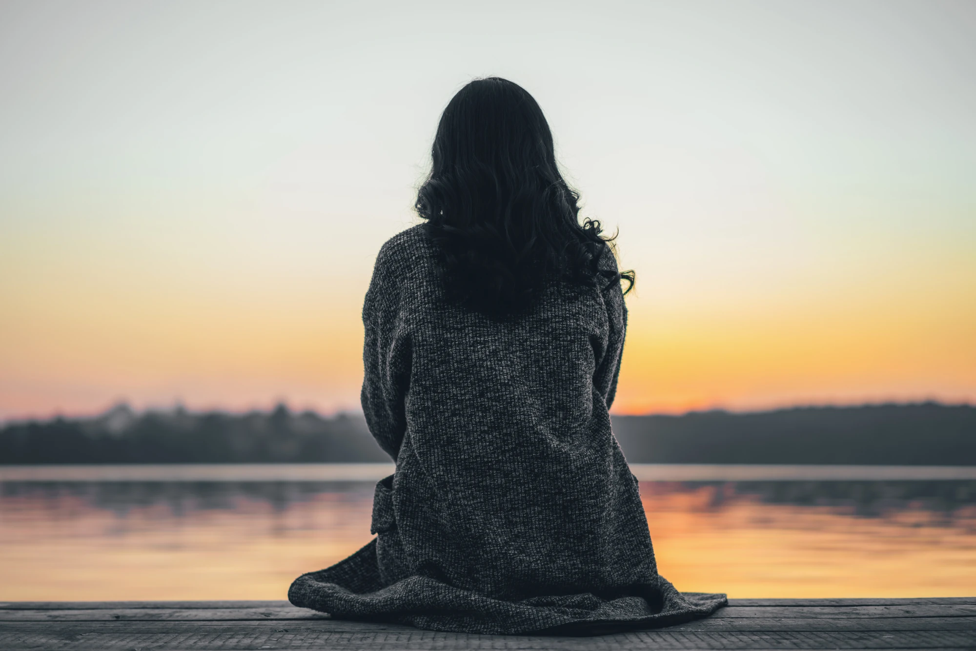  A woman sitting alone on a wooden dock by a calm lake, looking out at the water