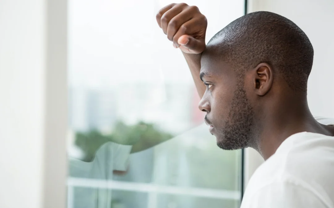 A man standing by a window at home, looking outside with a tired, thoughtful expression