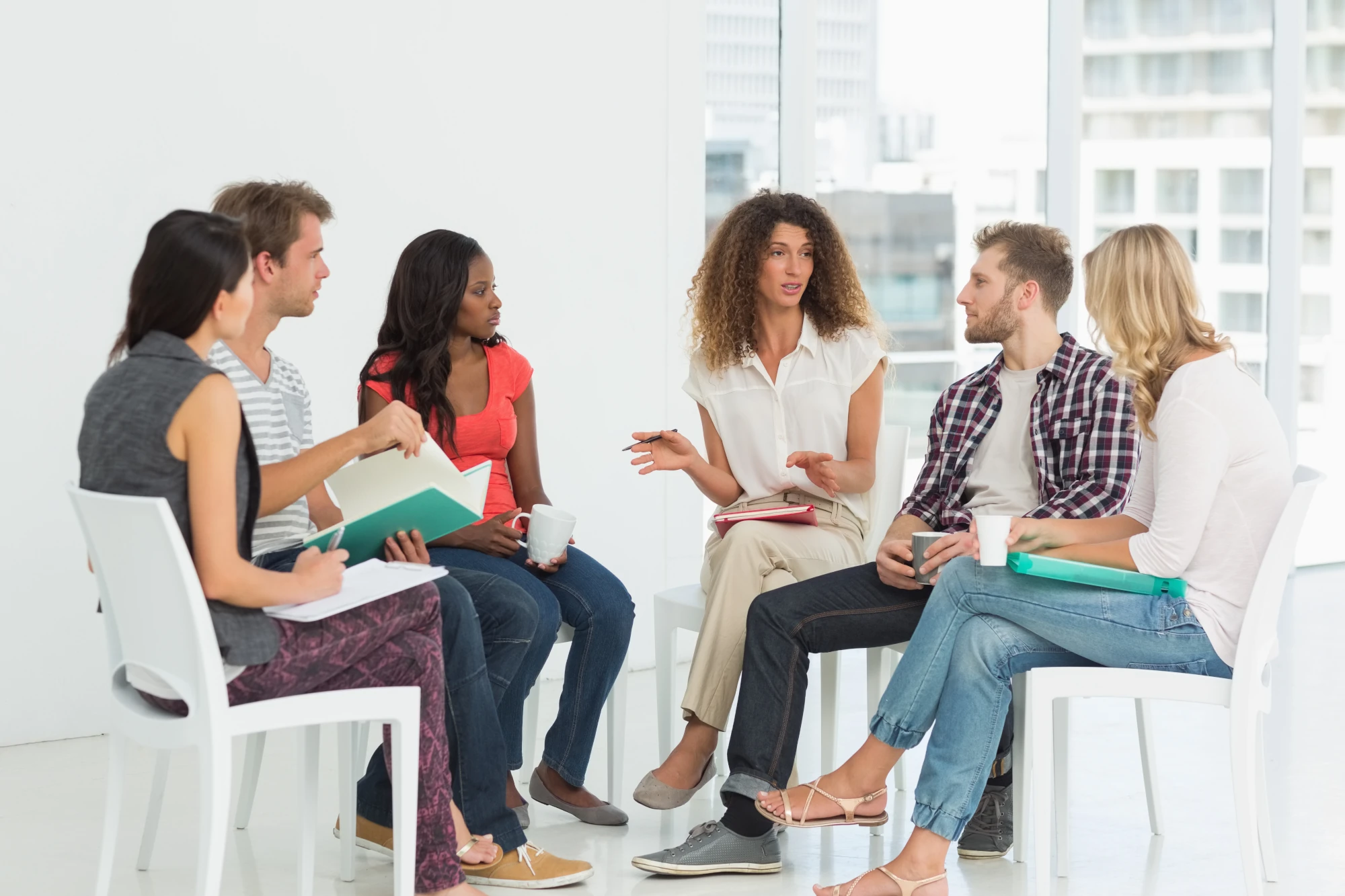 A small group of adults sitting in a circle of chairs in a bright room, talking with a counselor